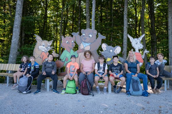 Eine Gruppe von Kindern und zwei Erwachsene sitzen auf Bänken vor großen Comicfiguren von Tieren in einem Wald.