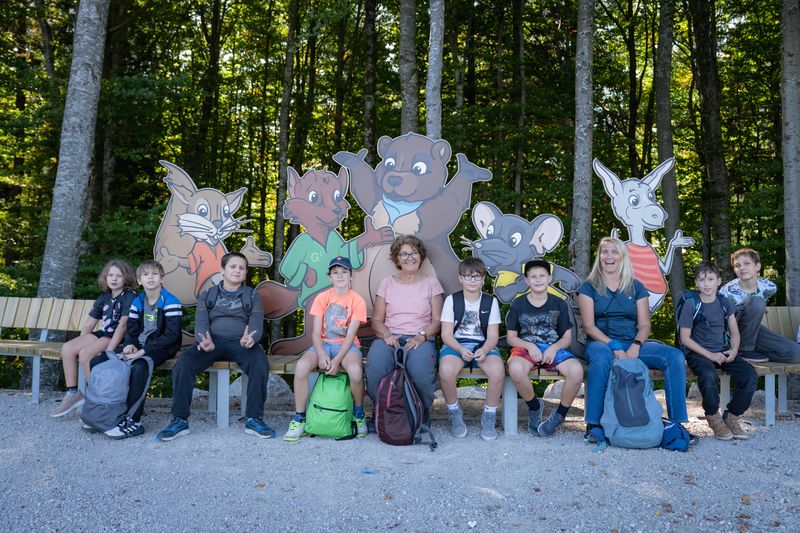 Eine Gruppe von Kindern und zwei Erwachsene sitzen auf Bänken vor großen Comicfiguren von Tieren in einem Wald.
