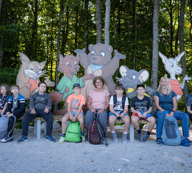 Eine Gruppe von Kindern und zwei Erwachsene sitzen auf Bänken vor großen Comicfiguren von Tieren in einem Wald.