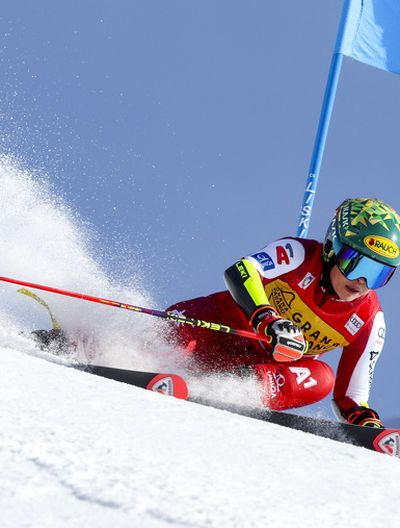 Skirennläuferin in rotem Anzug bei scharfer Kurve auf Skipiste, viel Schnee staubt auf. Dahinter steckt ein blaues Riesentorlauftor.