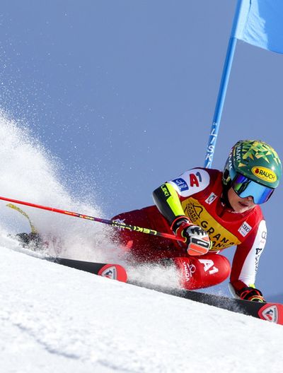 A female ski racer in a red suit makes a sharp turn on the ski slope, kicking up a lot of snow. Behind her is a blue giant slalom gate.
