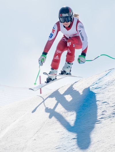 Skirennläuferin im roten Anzug springt über eine Schneekuppe. Die Sonne wirft lange Schatten auf die Piste neben einem roten Tor.