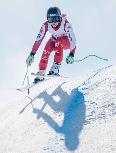 Skirennläuferin im roten Anzug springt über eine Schneekuppe. Die Sonne wirft lange Schatten auf die Piste neben einem roten Tor.