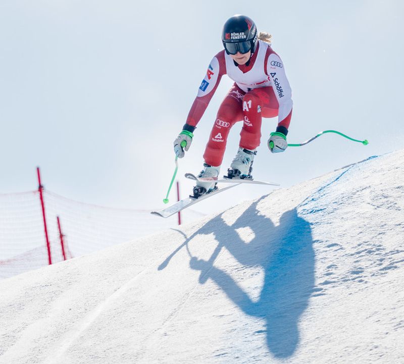 A female ski racer in a red suit jumps over a snow bump. The sun casts long shadows on the slope next to a red gate.