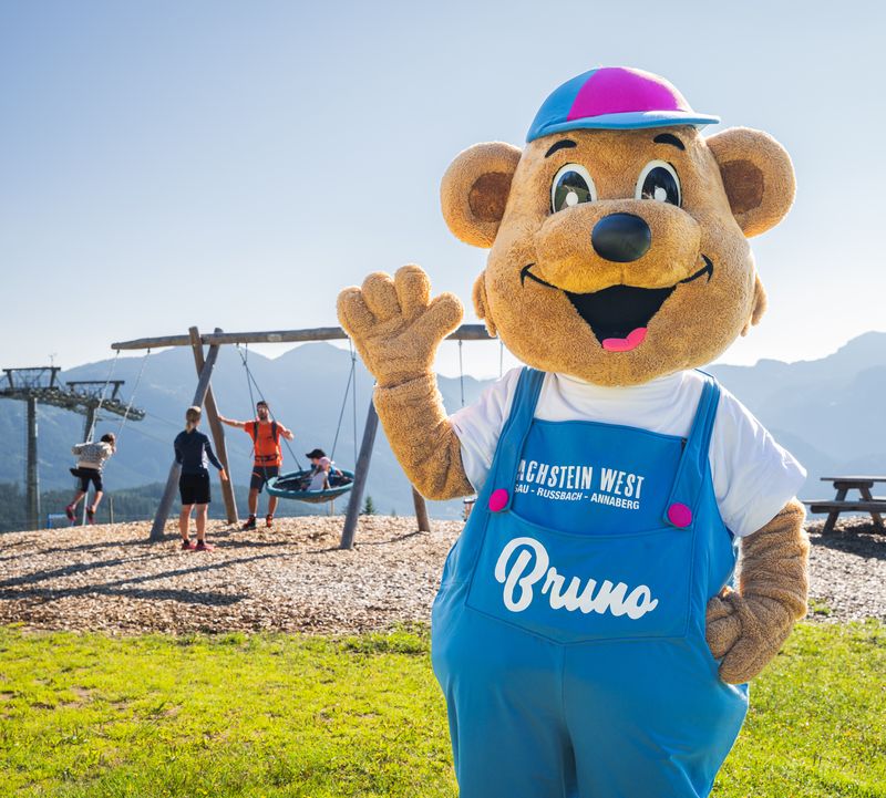 A bear mascot in a blue jumpsuit and cap waves. In the background, children swing on a playground against a picturesque mountain backdrop.