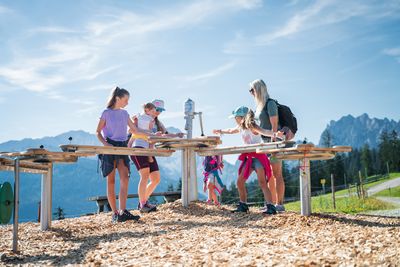 A group of people, including children, are playing at a wooden outdoor activity station in a mountainous area under a clear blue sky.