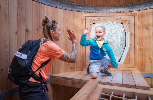 A happy woman and a boy are high-fiving inside a wooden play tower. The boy is laughing enthusiastically.