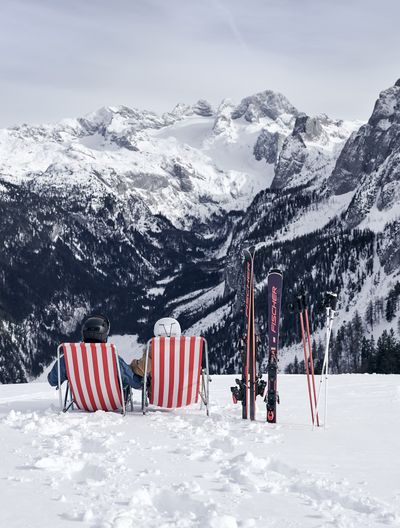 Zwei rot-weiß gestreifte Liegestühle stehen im Schnee, daneben sind zwei Paar Ski in den Schnee gesteckt. Dabei haben die Personen auf den Stühlen einen beeindruckenden Blick auf verschneite Gipfel und Wälder.