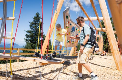 A father helps his son on a wooden and rope climbing course in the mountains in sunny weather.