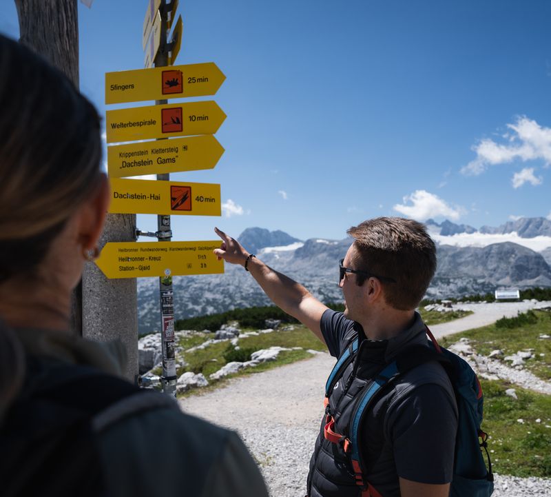 Wanderpaar studiert einen gelben Wegweiser in sonniger Berglandschaft mit schneebedeckten Gipfeln. Der Mann zeigt die Richtung.