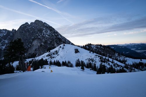 Pristine groomed ski slope with snow-covered hills, pine trees, and a rugged mountain peak under a clear blue sky.
