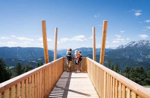A family on a wooden observation deck overlooking a vast mountain panorama under a blue sky.