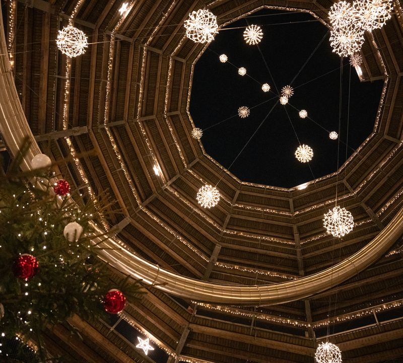 Interior shot of a round illuminated tower at night surrounded by brightly lit Christmas baubles and, on the left of the picture, the top of a decorated Christmas tree.