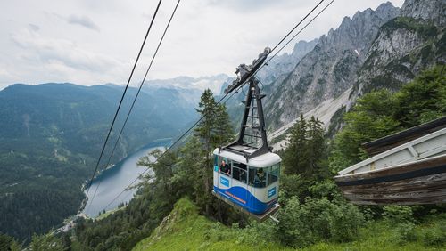 Bergbahn, die einen Berg hinauffährt, mit Blick auf den Gosausee und schroffe Gipfel und der schneebedeckte Dachstein Gletcher im Hintergrund, umgeben von üppigem Grün.