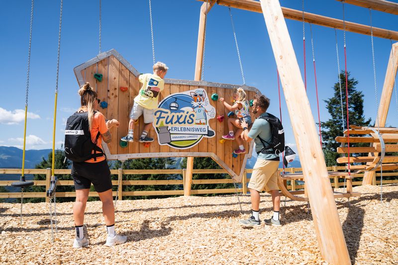 Familie am Spielplatz am Fuxis Erlebnisweg in Annaberg. Kinder klettern an der Kletterwand, Mama und Papa sehen ihnen zu und unterstützen sie.