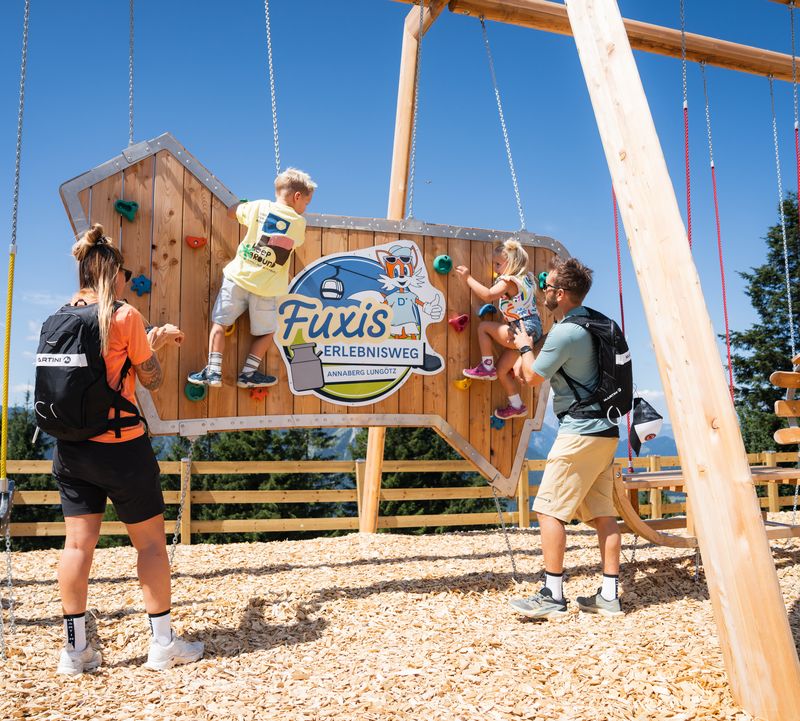 Familie am Spielplatz am Fuxis Erlebnisweg in Annaberg. Kinder klettern an der Kletterwand, Mama und Papa sehen ihnen zu und unterstützen sie.