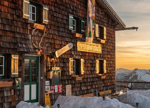 Rustic mountain hut with wooden shingles, snowy ground, and a sunset glow, adorned with antlers and a colorful sign outside – Christophorus Hut.