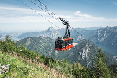Rote Gondel schwebt über bewaldetem Hang, im Hintergrund steile Berggipfel unter blauem Himmel.
