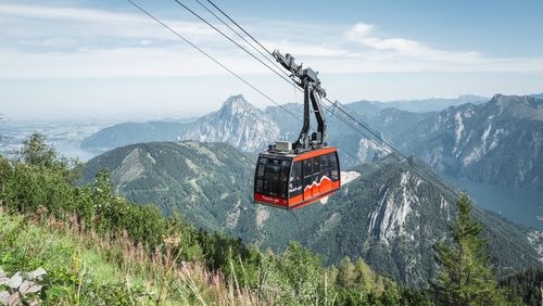 Rote Gondel schwebt über bewaldetem Hang, im Hintergrund steile Berggipfel unter blauem Himmel.