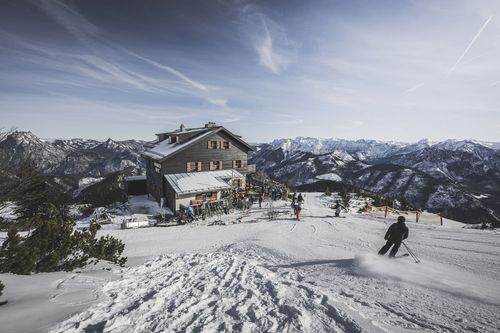 Skifahrer nahe einer Berghütte auf schneebedecktem Gipfel, im Hintergrund weiße Berge unter strahlend blauem Himmel – Hüttendorf Feuerkogel.