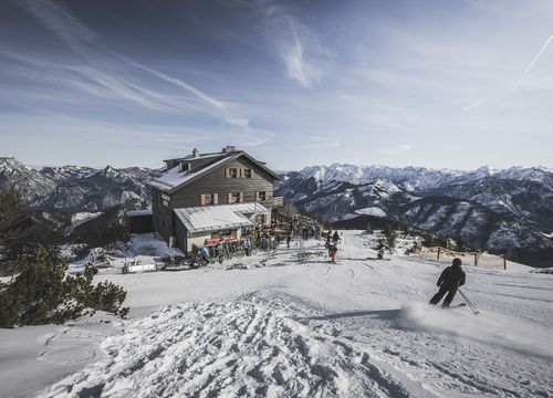 Skier near a mountain hut on a snow-covered summit, with white peaks in the background beneath a radiant blue sky – Kranabeth Hut, Feuerkogel.