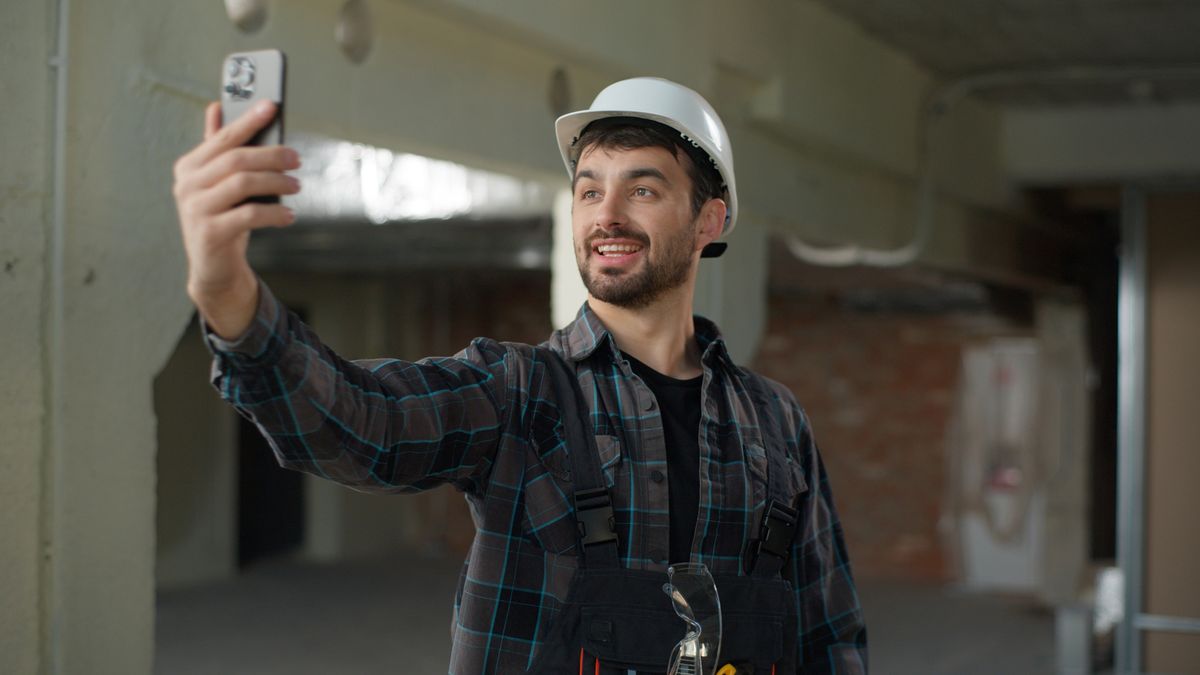 A construction worker in a hard hat takes a selfie inside an unfinished building, wearing a plaid shirt and overalls.