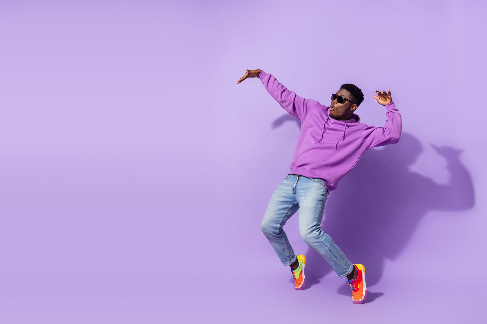 Person in a purple hoodie and jeans dancing energetically against a matching purple background, wearing sunglasses and bright orange sneakers.
