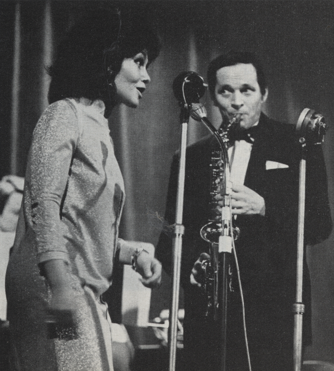 A woman sings into a microphone while a man plays the saxophone beside her on stage. Both are dressed formally.