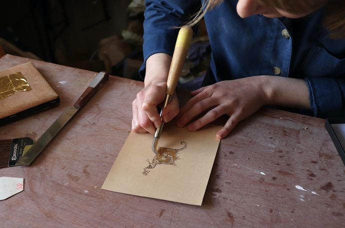 Person in a blue shirt using a carving tool to engrave a decorative design on a piece of paper at a wooden table.