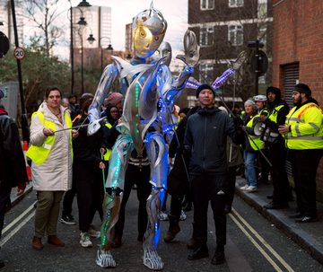 A towering metallic puppet with glowing blue and green lights is being guided through a street by event staff in high-visibility jackets during a parade.
