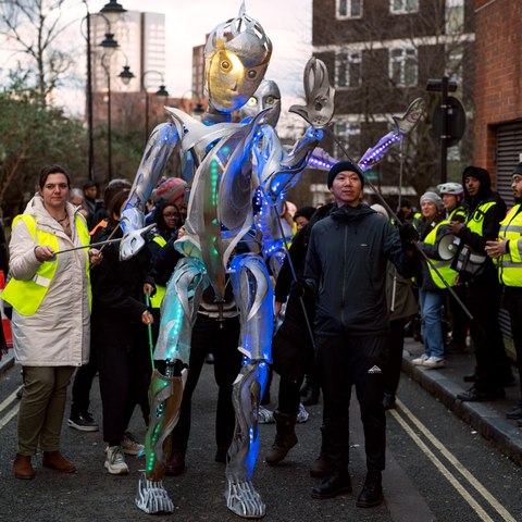 A towering metallic puppet with glowing blue and green lights is being guided through a street by event staff in high-visibility jackets during a parade.
