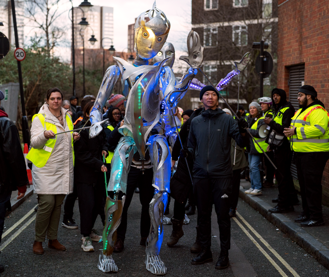 A towering metallic puppet with glowing blue and green lights is being guided through a street by event staff in high-visibility jackets during a parade.