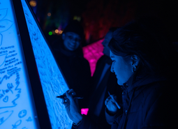 A person writes on a glowing blue board in a dark setting, surrounded by others. The scene is illuminated with vibrant purple and blue lights.