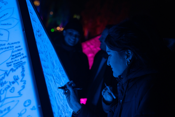 A person writes on a glowing blue board in a dark setting, surrounded by others. The scene is illuminated with vibrant purple and blue lights.