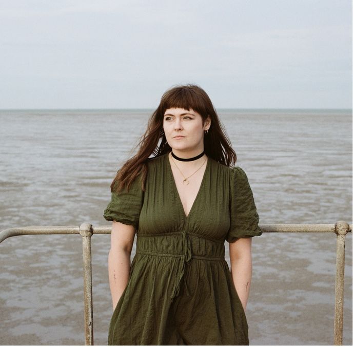 A person wearing an olive green dress and a black choker stands on a metal railing by the seaside with calm water and a cloudy sky in the background.