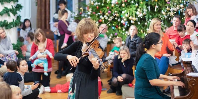 Musician playing violin and another on piano in front of a large, decorated Christmas tree, surrounded by a seated audience.
