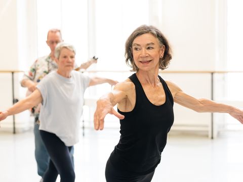 Elderly women and a man in a dance class, practicing ballet poses, smiling, with bright windows in the background.