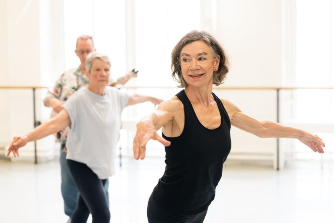 Elderly women and a man in a dance class, practicing ballet poses, smiling, with bright windows in the background.