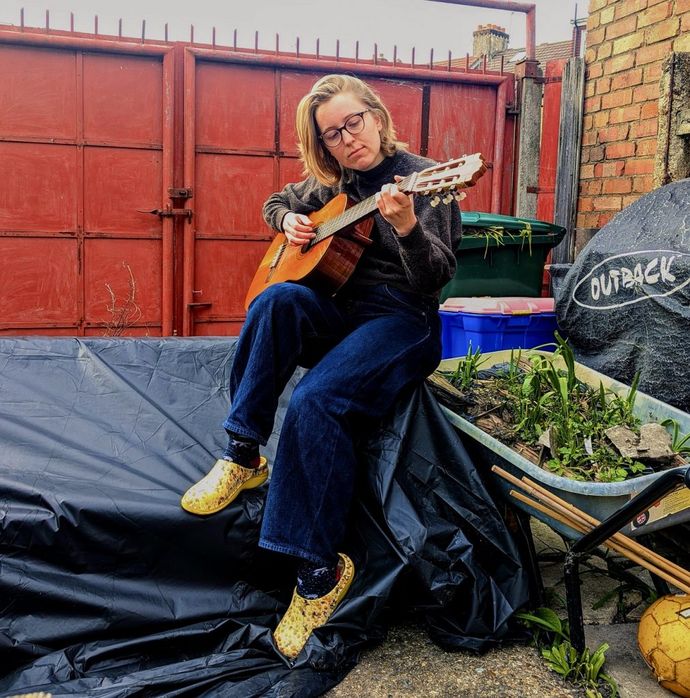 A person sits outdoors on a wheelbarrow covered with a tarp, playing an acoustic guitar in front of a red metal gate
