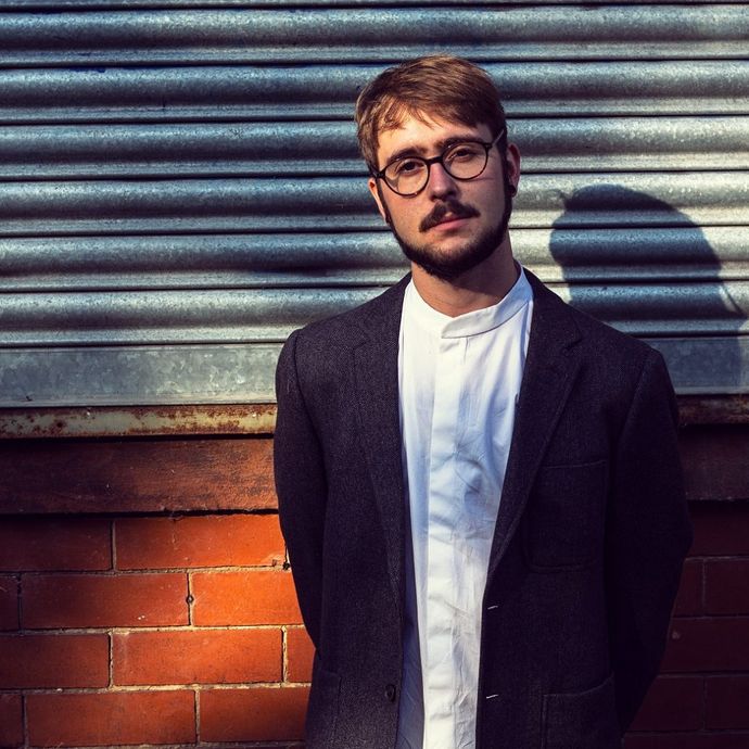 A man with glasses and a beard stands against a brick wall and metal shutter, wearing a dark blazer and white shirt, in sunlight.