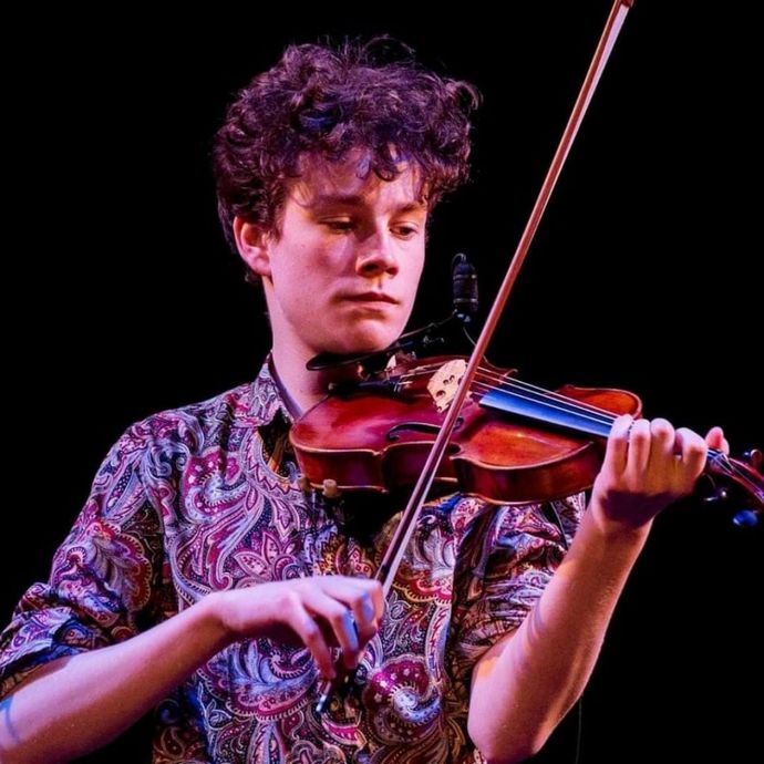 Person playing a violin on stage, wearing a colorful paisley shirt, focused expression, with a dark background.