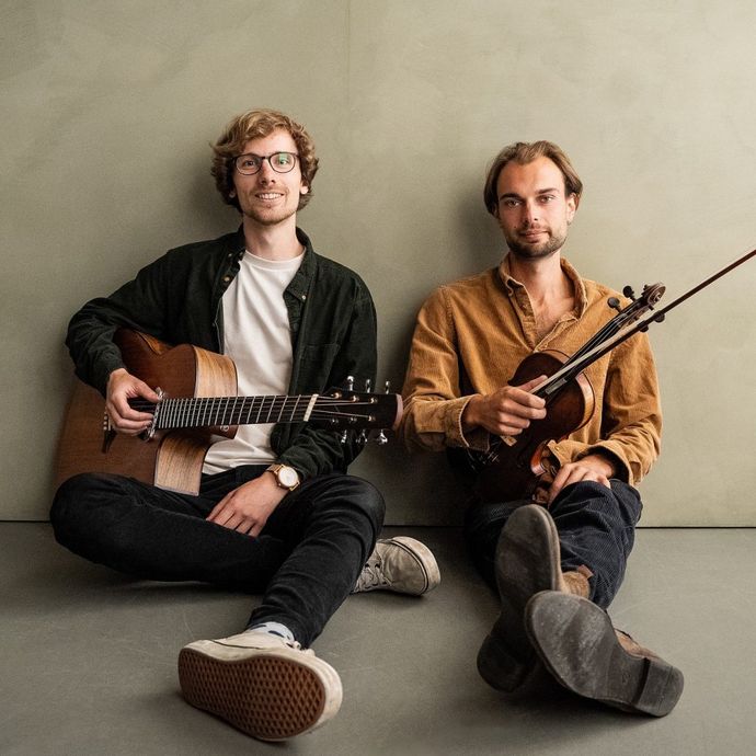 2 individuals sit on ground with a sandy background with 2 musical instruments in hand.