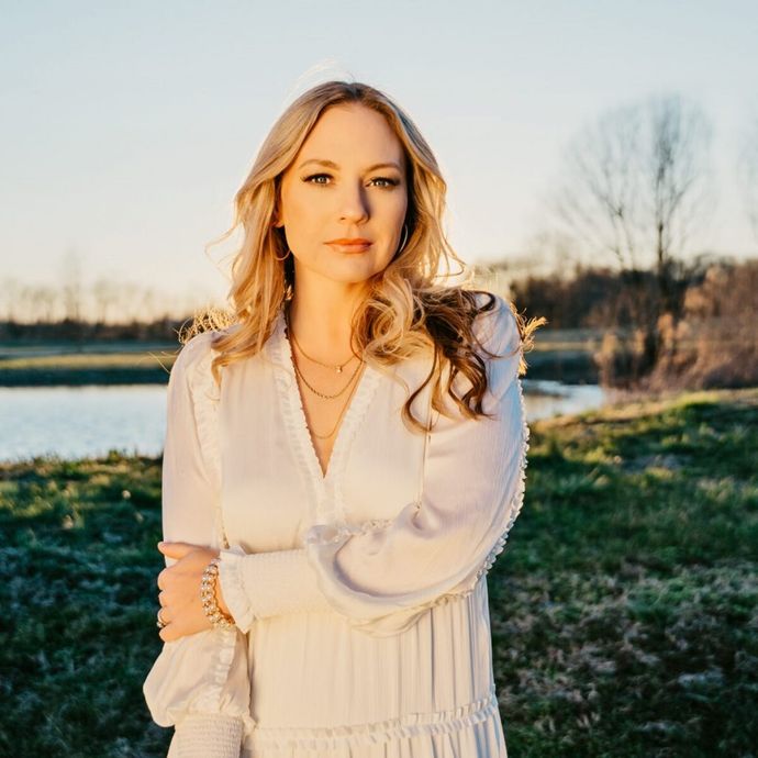Woman in a white dress standing by a pond at sunset, with long blonde hair and a serene expression, surrounded by a grassy landscape.