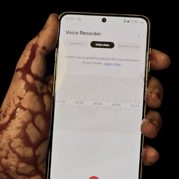 A phone held with a bloody hand with a voice recorder app shown to be open.