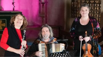 Three musicians performing indoors with instruments. One musician on the left is holding a clarinet and the musician in the centre is seated and playing an accordion, with a music stand in front. The musician on the right is standing and holding a violin and bow