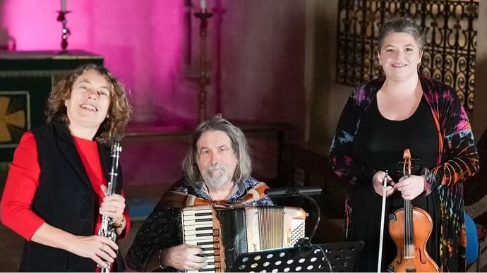 Three musicians performing indoors with instruments. One musician on the left is holding a clarinet and the musician in the centre is seated and playing an accordion, with a music stand in front. The musician on the right is standing and holding a violin and bow