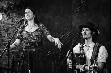 A woman sings with a microphone while a man plays guitar beside her in a dimly lit, plant-decorated setting. Both appear engaged and joyful.
