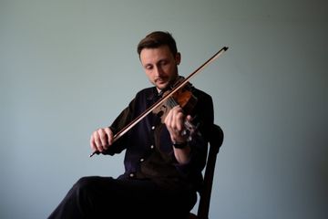 A musician seated, playing a violin against a plain light blue background, wearing a dark shirt and focused on his music.