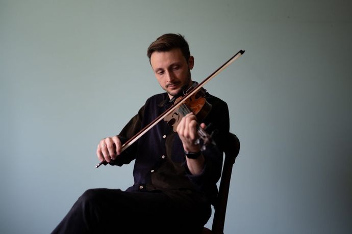 A musician seated, playing a violin against a plain light blue background, wearing a dark shirt and focused on his music.