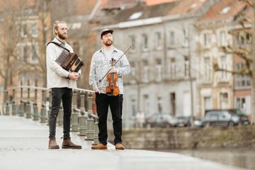 Two individuals stand on a bridge, one holding an accordion and the other a violin, with a blurred cityscape in the background.